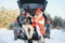 Couple in love sitting in car trunk drinking hot tea in snowy winter forest and chatting. People relaxing outdoors during road