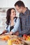 Couple making fresh organic juice in kitchen together. A young woman in a blue shirt slices a baguette. A man is hugging