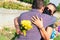 Couple with protective mourning mask holding flowers in hands and hugging at grave in cemetery. Tragic results of the coronavirus