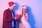 Couple in santa hats, new year, christmas. Lovers in neon light. Guy gives a gift in a red box to a happy girl, surprised woman in