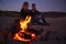 Couple Sitting On Surfboard By Camp Fire On Beach Using Mobile Phone As Sun Sets Behind Them