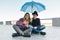 Couple of smiling teen friends sitting under an umbrella and looking at smartphone, lifestyle of teenagers in the city