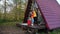 A couple with suitcases are hugging on the porch of a triangular wooden house.