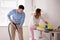 Couple Vacuuming Hardwood Floor And Dusting Desk