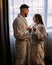 Couple wearing bathrobes in hotel standing near window with glasses of champagne