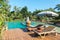 Couple of Wooden Chair under The Umbrella by The Swimming Pool surrounded by Various Type of Trees