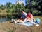 A couple of young people with gadgets are sitting on a blanket in a park near the lake. A man with a laptop and a woman with a