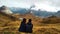 Couple of young travellers sitting on the grass looking to a valley with high mountains, lakes and clouds