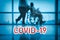 COVID-19 billboard red text on blue medical hospital background with doctor walking with disabled patient in wheelchair