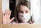 COVID-19 coronavirus concept, little girl in face mask looking through window at home or clinic. Portrait of sad kid during