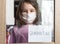 COVID-19 coronavirus concept, little girl in face mask looking through window with note Quarantine at home. Sad kid during