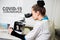 Covid-19 coronavirus poster. Young female doctor looking in a microscope in a clinic labalatory.