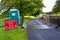 A Covid-19 social distancing warning sign and a plastic portable toilet next to a cattle grid on a public footpath at an outdoor