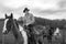 Cowboy on horseback, horse riding with chequered shirt with other horses, a gate, field and stone cottage in background