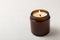 Cozy burning candle in brown glass jar on white texture wood.