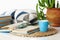 Cozy home interior decor: cup of coffee, stack of books, plants in pots, open book on a wicker stand, pillows on a white table.