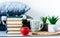 Cozy home interior decor: cup of coffee, stack of books, red apple, plants in pots on a wicker stand, blue pillows on a white