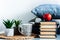 Cozy home interior decor: cup of coffee, stack of books, red apple, plants in pots on a wicker stand, blue pillows on a white