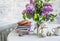 Cozy home still life - bouquet of lilacs, stack of books, glasses, toy bear, mug on the table in a bright room