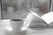 Cozy home still life: white cup of hot coffee and white opened book on windowsill against snow landscape outside