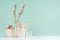 Cozy rustic home decor - beige dry flowers, plants in glass bottles, wicker basket, bunch twigs on green mint menthe wall, white.