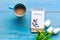 Cozy winter relax time.  Cup of hot coffee with write note on paper and lily flower on the blue wood background in morning sunny d