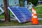 A cracked solar panel on the ground near a utility pole that it fell from with orange warning cones nearby