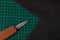 A craft knife lies on a green cutting Board, next to a piece of black leather