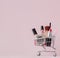 Creative concept with shopping trolley with makeup on a pink background. Perfume, sponge, brush, mascara, pencil, nail file, eye