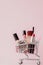 Creative concept with shopping trolley with makeup on a pink background. Perfume, sponge, brush, mascara, pencil, nail file, eye