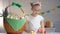 Creative girl in white dress showing painted Easter egg, decorated basket table