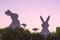 Creative photo of two silhouette paper rabbits in the chamomile flowers and green grass on the sunset sky background