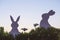 Creative photo of two silhouette paper rabbits in the chamomile flowers and green grass on the sunset sky background