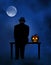 A creepy silhouetted man in a fedora with glowing eyes is seated next to a lighted jack-o-lantern