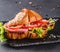 Croissant sandwich with prosciutto, tomatoes, cheese parmesan and greens on black shale board over black stone background. Healthy