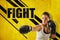 Crop close-up of young woman in boxing gloves holding one hand out, against yellow wall with `FIGHT` title and black