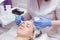 Crop master applying white mask with brush on face and neck of young woman in towel lying on table