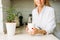 Crop photo of charming blonde woman with long fair hair in white shirt with morning cup of tea in hands on bright kitchen before