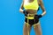 Crop shot of female bodybuilder with tape measure on waist on the blue background. Muscular body of a woman in sportswear with