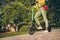 Cropped close up view photo of woman holding green eco friendly scooter with one foot on ground in park