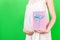 Cropped image of pink spotted gift box in pregnant woman`s hand against her belly at green background. Future mother in white