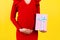 Cropped image of pink spotted gift box in pregnant woman`s hand against her belly at yellow background. Future mother in red dres