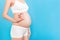 Cropped image of positive pregnant woman in white underwear showing okay gesture against her belly at blue background. Easy and