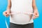 Cropped image of pregnant woman in home clothing measuring her growing abdomen at blue background. Inch tape measure. Copy space