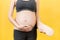 Cropped image of pregnant woman on the third trimester putting on pregnancy belt at yellow background. Orthopedic abdominal