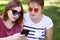 Cropped outdoor shot of two female best friends in trendy shades, dressed casually, read information via smart phone in internet,