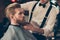 Cropped photo of a classy dressed barber shop stylist working for a perfect look of a red bearded guy in a cape. His hairdo looks