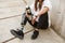 Cropped photo of handicapped woman having bionic leg in streetwear, sitting on concrete floor outdoor and holding thermos cup