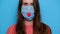 Cropped portrait of cute young woman, isolated on blue wall, wearing health mask with little red heart