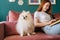 Cropped shot of cheerful young woman reading book lying on cozy sofa with pretty miniature spitz dog in living room with